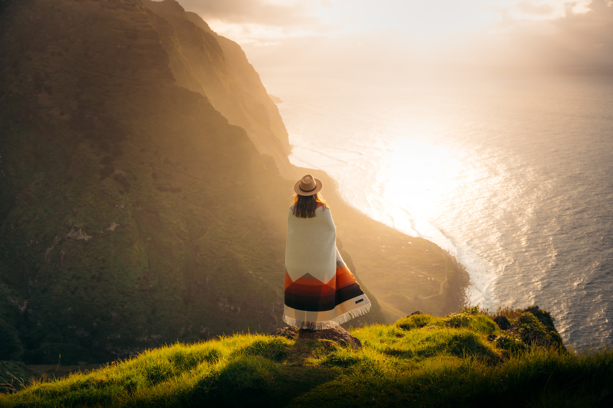 Person wrapped in a Sackcloth & Ashes blanket on coastal cliffs at golden hour